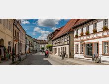 Gepflasterte Altstadtstraße mit historischen Fachwerkhäusern und Gebäuden im klassischen Stil, teilweise mit Blumenkästen an den Fenstern. Menschen gehen und fahren Fahrrad entlang der Straße, während einige Geschäfte und Auslagen sichtbar sind; der Himmel ist blau mit weißen Wolken.