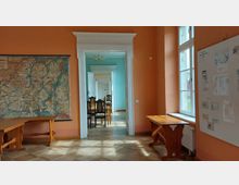 Ein Innenraum mit orangefarbenen Wänden und einer großen Karte an der linken Wand. Durch eine türgerahmte Sichtachse sind dahinterliegende, hellblaue Räume mit Holzmöbeln und einem Kronleuchter sichtbar. Ein großes Fenster auf der rechten Seite lässt Tageslicht in den Raum.