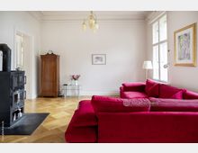 Wohnzimmer mit hellem Holzboden, einer großen roten Eckcouch, einem schwarzen Kaminofen auf einer schwarzen Matte und einem Holzschrank an der Wand. Der Raum ist mit weißen Wänden, hohen Fenstern und einem modernen Kronleuchter gestaltet.