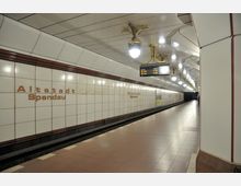 U-Bahn-Station "Altstadt Spandau" in Berlin mit hellen Wandfliesen, rot-braunen Akzenten und einer hohen Decke mit dekorativen Lampen. Im Vordergrund sind ein Bahnsteig und eine elektronische Infotafel sichtbar.