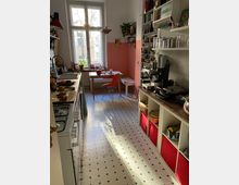 Schmale Küche mit großen Fenstern, die Tageslicht hereinlassen. Links ein Gasherd und Spüle, rechts Regale mit Geschirr, einer Kaffeemaschine und roten Aufbewahrungsboxen; am Ende steht ein kleiner Tisch mit zwei Stühlen, die Wand ist teils rosa gestrichen.