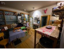 Gemütlicher Innenraum eines kleinen Cafés oder Ladens mit Holzfußboden, Vitrinen und Regalen voller Flaschen und Gläser sowie einem Tisch mit blauem Tischtuch und bunten Stühlen. Im Vordergrund steht eine hohe Holztheke mit rot-weiß kariertem Läufer, an der Wand hängen Dekorationen und Regalbretter mit weiteren Produkten.