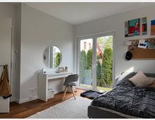 Bright modern bedroom with white walls, a low bed, and a small vanity desk with a round mirror and chair. Large glass patio doors open to a view of tall evergreen shrubs, and a corkboard with photos hangs on the wall.