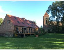 Backsteinbau mit rotem Satteldach und Holzbalkon, umgeben von einem Garten mit Blumen, auf einer großen Rasenfläche. Im Hintergrund ein historischer Kirchturm aus Stein mit rotem Ziegelwerk, flankiert von hohen Bäumen.