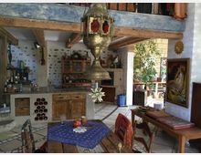 Wohnraum mit rustikaler Einrichtung, bestehend aus einer offenen Küche mit Holzschränken, freiliegenden Holzbalken und einem Esstisch aus Holz im Vordergrund. Der Raum enthält dekorative Details wie eine orientalische Metalllampe, ein Kunstgemälde, ein Bücherregal und eine Pflanze in einer Ecke.