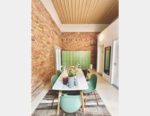 Ein modern eingerichteter Besprechungsraum mit einer langen weißen Tischplatte und grünen Stühlen, umgeben von Wänden aus sichtbaren Ziegelsteinen. Im Hintergrund stehen grüne Schließfächer, und auf dem Tisch befindet sich eine Blumenvase mit bunten Blumen.