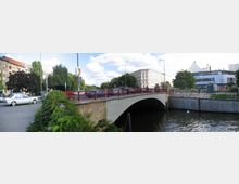 Straßenbrücke über einen Fluss mit roten Geländern, flankiert von Grünflächen und Gebäuden im Hintergrund, darunter ein mehrstöckiges Wohnhaus und ein modernes Gebäude mit Schildern. Autos parken auf der Straße, und die Uferbereiche sind mit Pflastersteinen und Vegetation gestaltet.