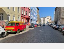 Eine gepflasterte Straßenszene in einem Wohngebiet mit mehrstöckigen Häusern in verschiedenen Farben (hellgelb, rot, blau und braun) auf der linken Seite. Am Straßenrand steht ein bunt lackierter, gelb-roter Kleinbus zwischen anderen geparkten Autos, während die Straße weiter in ein moderneres Gebäude mündet.