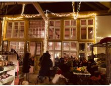 Innenansicht eines gemütlichen, rustikalen Gebäudes mit großen Fenstern und Holzelementen, geschmückt mit Lichterketten. Menschen sitzen an einem Tisch, während andere vorbeigehen; links im Vordergrund sind Regalwaren wie Gläser mit Marmelade zu sehen.