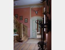 Holzvertäfelter Eingangsbereich mit einer geschwungenen Treppe, roten Wänden und Holzboden. Über einer offenen Tür mit grünen Verzierungen hängt ein Geweih, daneben sind gerahmte Bilder an der Wand und ein Kleiderständer steht im Vordergrund.