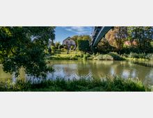 Eine ländliche Landschaft mit einem Fluss im Vordergrund, gesäumt von Bäumen und Gras. Im Hintergrund steht ein Haus mit rotem Dach, umgeben von einem Garten, und darüber führt eine Brücke.
