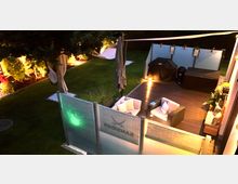 Beleuchtete Terrasse mit Holzboden, Gartenmöbeln und Grill in einem privaten Garten. Im Hintergrund ist eine gepflegte Rasenfläche mit einer Hängematte unter Bäumen zu sehen, die von Pflanzen und Dekorationsleuchten umrahmt ist.