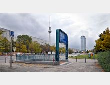 Straßenansicht in Berlin mit dem Eingang zur U-Bahn-Station „Schillingstraße“ im Vordergrund, einem hohen blauen Stationsschild und Metallgeländern. Im Hintergrund sind der Berliner Fernsehturm, moderne Gebäude und herbstlich gefärbte Bäume zu sehen.