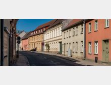 Curving residential street lined with closely spaced, two- to three-story pastel stucco row houses with red-tiled roofs and white-framed windows. Narrow sidewalks, street signs, and utility pipes run along the building facades under a clear blue sky.