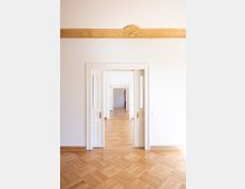 Ein heller Innenraum mit Fischgrät-Holzparkett, weißen Wänden und mehrfach hintereinander angeordneten weißen Flügeltüren mit Glaseinsätzen. Über der Türöffnung verläuft ein dekoratives, goldfarbenes Zierband mit floralen Ornamenten.