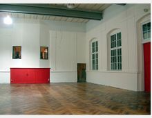 Ein leerer Innenraum mit Parkettboden, weißen Wänden und großen Fenstern mit Sprossen. An der Wand befinden sich zwei kleine Fenster und eine rote Holztür sowie rote Holzelemente als Akzent.