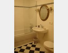 Ein Badezimmer mit weiß gefliesten Wänden und schwarzen Mosaikbordüren. Im Vordergrund befindet sich ein Waschbecken mit ovalem Spiegel in einem goldfarbenen Rahmen an der Wand, daneben zwei Wandleuchter. Der Boden ist schwarz-weiß kariert, und eine Badewanne mit einer Glasduschwand ist im Hintergrund sichtbar.