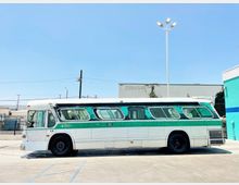 Ein älterer Linienbus mit grün-weißer Lackierung steht auf einem Parkplatz bei Tageslicht. Im Hintergrund sind ein Gebäude mit Industriecharakter, ein Zaun und Straßenbeleuchtung zu sehen.