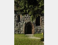 Historische Kirchenfassade aus unregelmäßigem Natursteinmauerwerk mit gotischem Spitzbogenportal, flankiert von zwei hohen, schmalen Fenstern mit Bogenabschluss. Im Vordergrund befindet sich eine Wiese, teils von überhängenden Tannenzweigen beschattet.