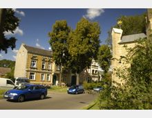 Straßenszene mit Wohnhäusern aus gelbem und rotem Backstein neben Bäumen; rechts im Bild ein Teil einer Kirche mit gelbem Mauerwerk und spitzem Turm, der mit einem goldenen Stern verziert ist. Im Vordergrund parken Autos entlang der Straße.