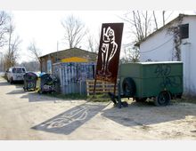 Außengelände mit Schotterweg, grauen Müllcontainern und einem rostfarbenen Metallkunstwerk mit ausgeschnittenem Fischmotiv. Im Hintergrund stehen ein altes Ziegelgebäude, ein grüner Anhänger und weitere mit Graffiti bedeckte Metallstrukturen, umgeben von kahlen Bäumen.