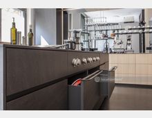 Moderne Küche mit dunkler Kücheninsel und integrierten Edelstahlschubladen, daneben ein Spülbereich mit Armatur. Im Hintergrund helle Schränke, Hängeregale mit Gläsern und Tassen sowie eine Kaffeemaschine.