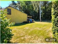 Gelbes Haus mit Satteldach am Rand einer Wiese, umgeben von Bäumen und Sträuchern; im Hintergrund dichter Wald.
