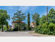 Straßenszene in einem Wohngebiet mit Kopfsteinpflaster, umgeben von Bäumen und Büschen. Im Hintergrund stehen Einfamilienhäuser mit roten Ziegeldächern, teils verdeckt durch hohe Nadel- und Laubbäume.