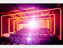 Kleiner Kinosaal mit mehreren Reihen gepolsterter Sitze in dunklem Blau. Hell leuchtende Neonlichter in Orange und Rot rahmen die Wände ein, während ein Projektor ein starkes Licht auf die Leinwand projiziert.