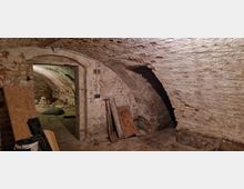 Gewölbekeller mit unverputzten Ziegelwänden und einem Rundbogen, der in einen weiteren Raum führt. Der Boden besteht aus groben Ziegelsteinen, und im Vordergrund liegen Holzplatten und Baumaterialien.