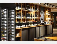 Innenansicht eines Weinladens mit langen Holzregalen voller Weinflaschen entlang der Wand und einer Theke davor. Links stehen beleuchtete Weinkühlschränke mit Glasfront, darüber hängen schwarze Schienenstrahler an der Decke.