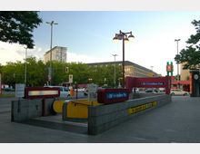 Eingang zum U-Bahnhof Fehrbelliner Platz in Berlin mit rotem und gelbem Design, umgeben von Bäumen, Straßenverkehr und einem hohen Gebäude im Hintergrund. Eine Uhr und Straßenbeleuchtung sind sichtbar, der Bereich vermittelt eine städtische Szenerie.