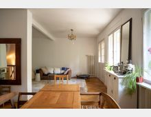 Heller Wohn- und Essbereich mit Holztisch im Vordergrund, Sitzbank und Teppich im Hintergrund sowie Parkettboden und Kronleuchter an der Decke. Rechts sorgen große Fenster und eine verglaste Tür für viel Tageslicht; darunter steht ein Sideboard mit Pflanzen und Dekoration, daneben ein hoher Heizkörper.