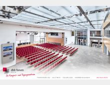 Großer moderner Konferenzsaal mit einer Bestuhlung aus roten Stühlen, einer Bühne mit Podium, großen Fenstern, Holzverkleidungen und einer hellen, offenen Decke mit Beleuchtungselementen. Im Hintergrund ist ein Eingangsbereich mit Glasfront und Treppen sichtbar.