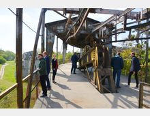 Eine alte, verrrostete Stahlkonstruktion, wahrscheinlich Teil einer historischen Industrieanlage, mit offenen Metallträgern und mechanischen Teilen. Mehrere Personen stehen oder gehen entlang des Betonbodens, während grüne Vegetation und ein Gebäude im Hintergrund sichtbar sind.