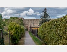 Schmaler gepflasterter Weg zwischen hohen Hecken, der zu einer historischen Steinmauer mit einem gemauerten Rundbogeneingang führt; im Hintergrund ein einzelner Nadelbaum und bewölkter Himmel.