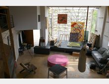 Offenes Kunststudio mit hoher Decke und großen Fenstern, durch die Tageslicht einfällt. Im Raum befinden sich Staffeleien, abstrakte Gemälde, ein Boxsack, eine graue Couch, Holzstapel und diverse Arbeitsmaterialien, die eine kreative Arbeitsumgebung schaffen.