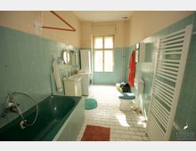 Großes Badezimmer mit hellgrünen Wandfliesen, einer dunklen Badewanne, einem Waschbecken mit Spiegel, einer Toilette und einem Handtuchheizkörper. Der Raum hat ein Fenster, viel Tageslicht und geflieste Böden, sowie eine Waschmaschine und einen Wäschekorb.