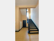 Treppenhaus in einem Gebäude mit hellbeigen Wänden und dunklem Treppengeländer. Im Hintergrund befindet sich ein großes, halbkreisförmiges Fenster mit Blick auf die obere Etage.