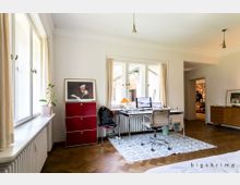 Helles Arbeitszimmer mit großen Fenstern, einem Schreibtisch mit zwei Monitoren, einer Lampe und einem Bürostuhl auf einem gemusterten Teppich. Im Hintergrund sind weitere Räume und Bücherregale sichtbar, der Boden besteht aus Parkett.