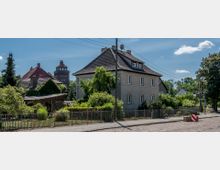 Wohngebiet mit einem zweistöckigen Haus in grauer Putzfassade und Satteldach, umgeben von üppigem Grün und einem Zaun. Im Hintergrund ist ein roter Backsteinturm mit Fenstern und einer geschwungenen Dachspitze zu sehen.