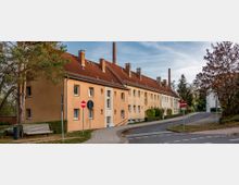 Mehrfamilienhaus in einer Wohnsiedlung mit gelblich-beiger Fassade und roten Ziegeldächern, ergänzt durch mehrere kleine Schornsteine. Im Vordergrund befinden sich ein Gehweg mit Parkbank, Bäume sowie Straßenschilder, darunter ein Durchfahrtsverbot.