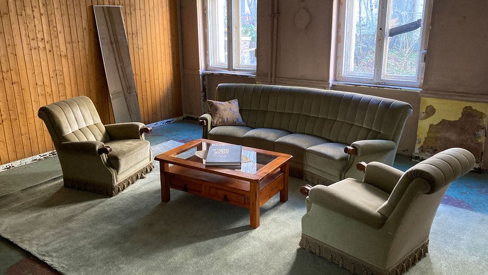 Wohnzimmer in einem teils renovierungsbedürftigen Altbau mit großen Sprossenfenstern, Holzvertäfelung an einer Wand und abblätterndem Putz. In der Mitte steht eine Sitzgruppe aus grünem Sofa und zwei Sesseln um einen niedrigen Holztisch auf einem großen Teppich.