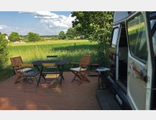 Wohnmobil mit geöffneter Tür, geparkt auf einer Terrasse mit Holzdielen. Darauf stehen ein Tisch mit Stühlen und ein Grill; im Hintergrund eine grüne Wiese mit Bäumen und vereinzelten Häusern unter einem blauen Himmel.