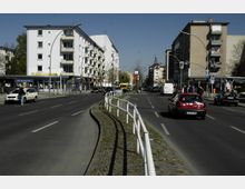 Städtische Straßenszene mit mehrspuriger Fahrbahn, flankiert von Mehrfamilienhäusern und Geschäften; eine Berliner Bank und der Eingang zur U-Bahn-Station "Bayerischer Platz" sind auf der linken Seite sichtbar. Im Vordergrund verläuft eine mit weißen Geländern abgetrennte Mittelinsel, Autos und Fußgänger beleben die Szene.