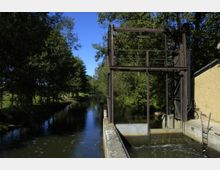 Metallische Schleusenanlage an einem schmalen Fluss, umgeben von dichtem Grün und Bäumen, mit einem gelblichen Gebäudeteil am rechten Bildrand und klarem blauen Himmel im Hintergrund.