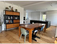 Wohnküche mit modernem Design: Im Vordergrund ein großer Holztisch mit bunten Stühlen auf Parkettboden im Fischgrätmuster. Im Hintergrund eine weiße Einbauküche mit schwarzer Kücheninsel, daneben ein offenes Regal aus Holz und Metall mit Dekorationsartikeln.