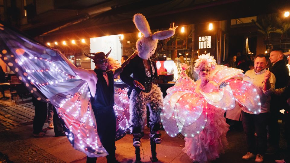 Nächtliche Außenansicht eines überdachten Bar- und Veranstaltungsbereichs mit warmen Lichterketten, Pflasterboden und dem Leuchtschild „BAR 100“. Im Vordergrund sind dekorative, bunt beleuchtete Kostüm- und Flügelelemente zu sehen, im Hintergrund stehen Tische und Eingangsbereiche unter dem Vordach.