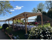 Holzüberdachter Außenbereich eines Restaurants mit gedeckten Tischen, direkt am Ufer eines Sees. Im Hintergrund stehen weitere Tische und Stühle auf einer grünen Wiese, umgeben von Bäumen und Pflanzen.
