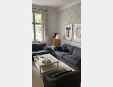 Wohnzimmer mit zwei grauen Sofas und einem grauen Sessel, einer weißen Wand mit geblümter Tapete und zwei gerahmten Kunstwerken. In der Mitte steht ein weißer Couchtisch mit Kerzenhaltern und Gläsern, daneben ein Fenster mit weißen Vorhängen, das Tageslicht einlässt.
